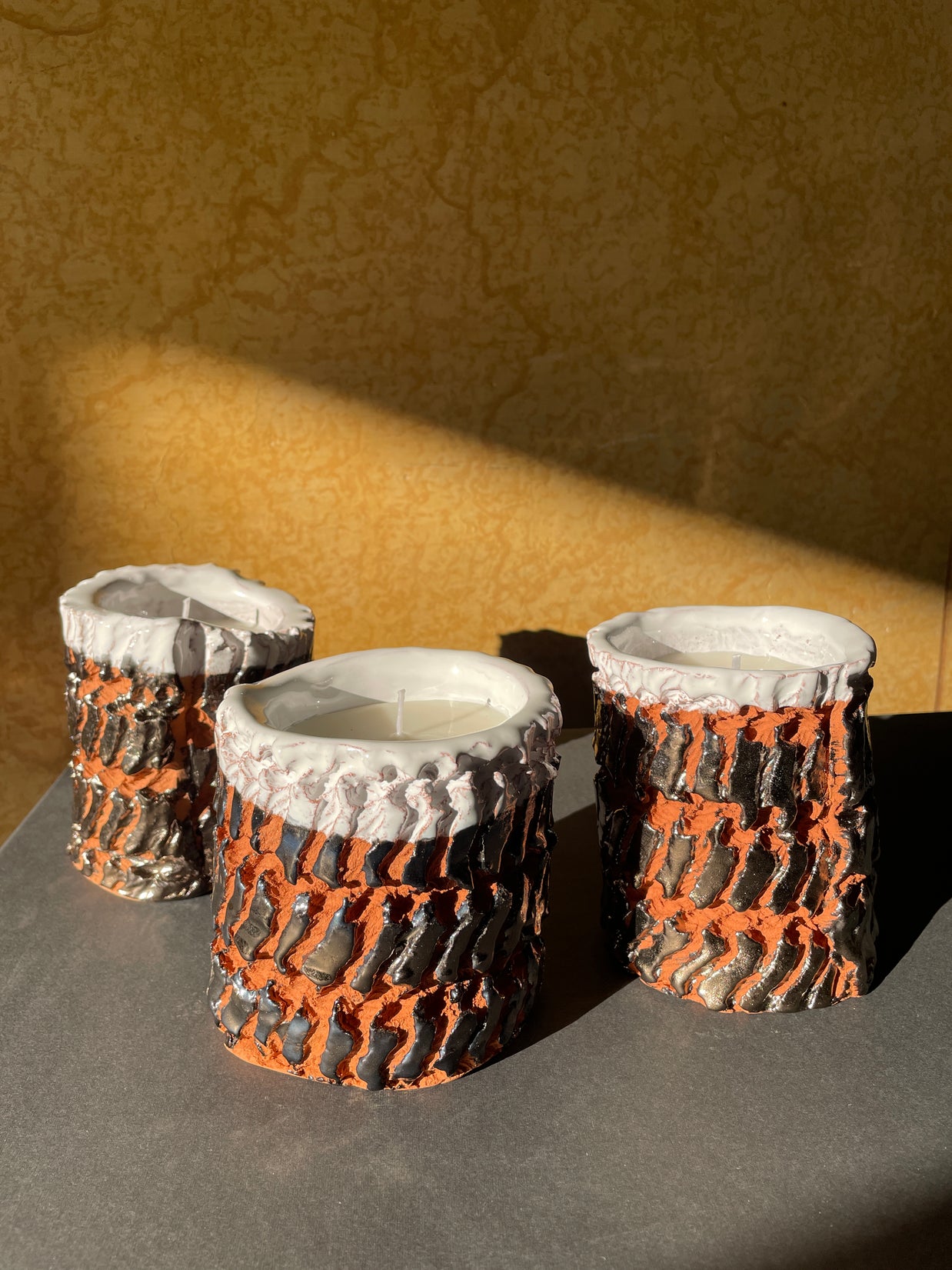 Three Ceramic Pot Candles in platinum glaze displayed on a taupe surface with a yellow wall backdrop, creating a warm and sophisticated composition.