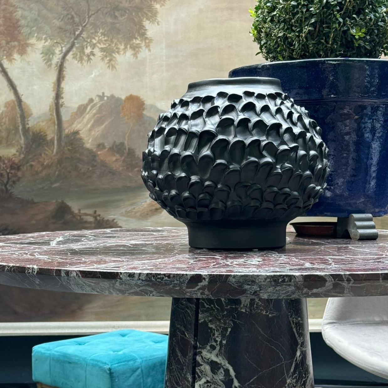 Indoor setting featuring the Ceramic Vase in Licorice Black on a marble table, accompanied by a blue vase in the background, a vibrant turquoise velvet stool, and a framed artwork.