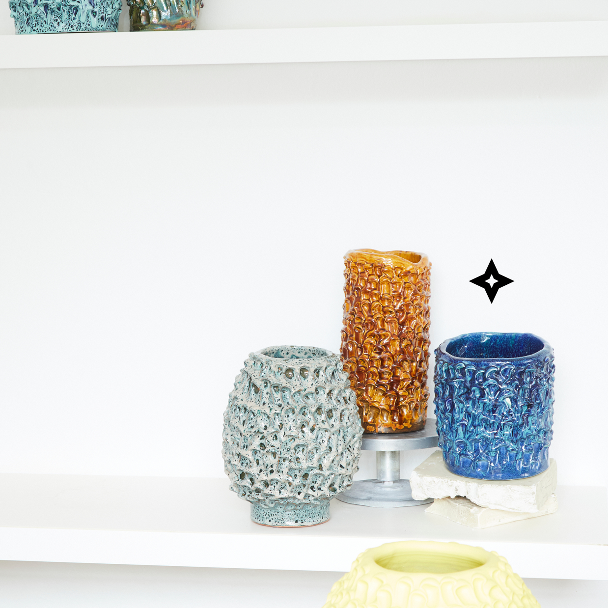 Display of three vases including the Whispy Matte Blue and Liquid Bronze Ceramic Vase, with complementary colors. One vase on a silver pedestal, all arranged on a white surface and against a white background.