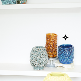 Display of three vases including the Whispy Matte Blue and Liquid Bronze Ceramic Vase, with complementary colors. One vase on a silver pedestal, all arranged on a white surface and against a white background.