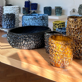 Composition of multiple Ceramic Vases in various colors, displayed on wooden shelves and tables at different heights, creating a visually dynamic arrangement.